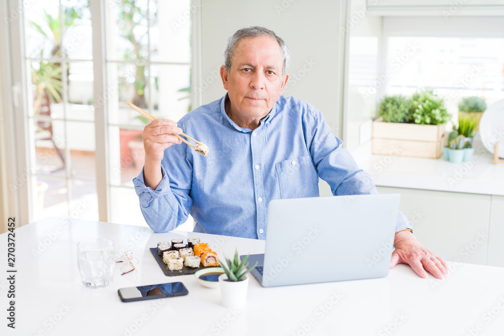 Handsome business senior man eating delivery sushi while working using laptop with a confident expression on smart face thinking serious
