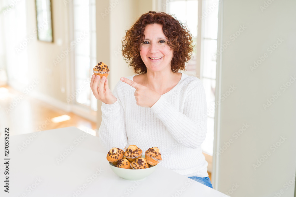 Senior woman eating chocolate chips muffins very happy pointing with hand and finger Senior woman eating chocolate chips muffins very happy pointing with hand and finger