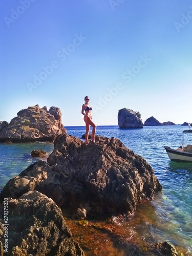 Attractive young woman posing in a black swimsuit on the cliff of the tropical island with the boat near