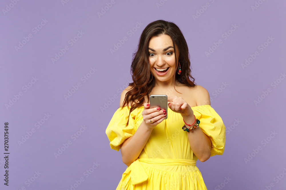 Surprised young woman in yellow summer dress using mobile phone, typing sms message isolated on pastel violet wall background in studio. People sincere emotions, lifestyle concept. Mock up copy space. Surprised young woman in yellow summer dress using mobile phone, typing sms message isolated on pastel violet wall background in studio. People sincere emotions, lifestyle concept. Mock up copy space.