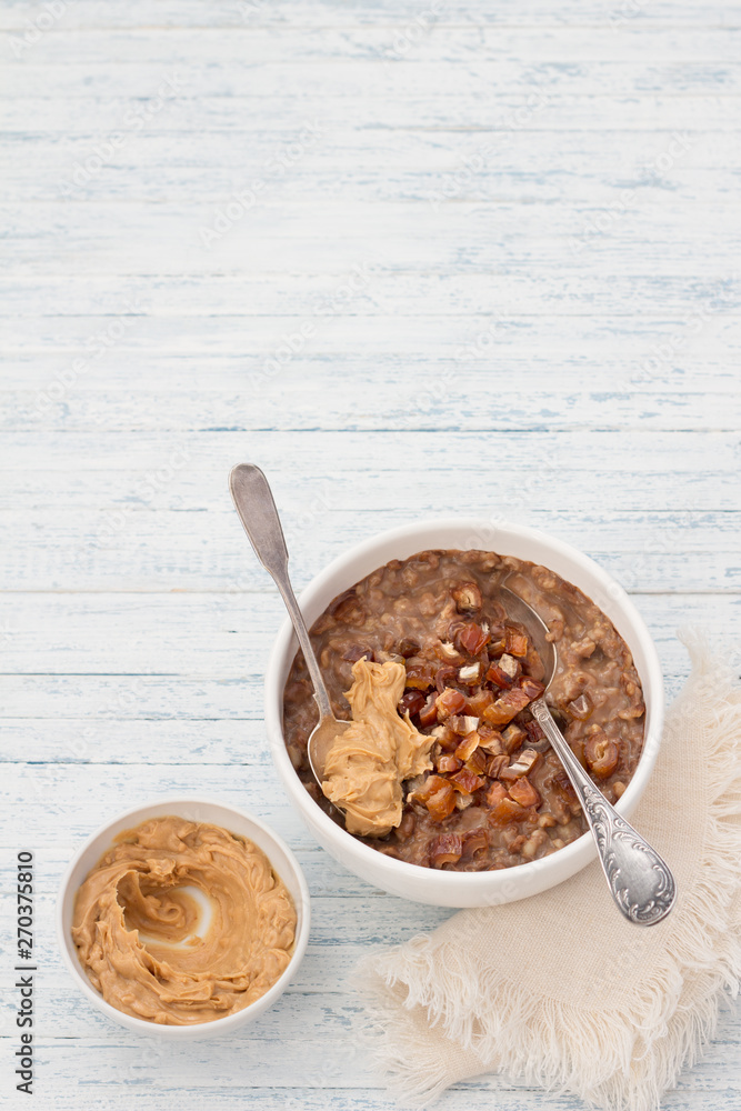 Fototapeta premium Delicious oatmeal in a white bowl, dates fruits, cocoa and vegan milk. healthy breakfast on light blue background. top view. free space