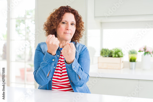 Middle age senior woman with curly hair wearing denim jacket at home Ready to fight with fist defense gesture  angry and upset face  afraid of problem
