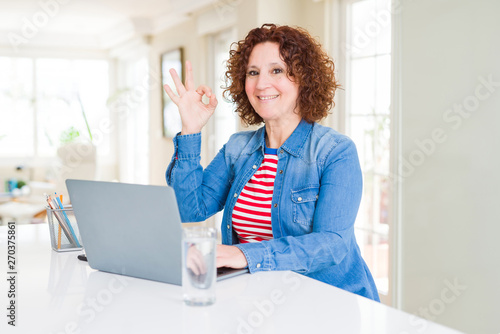 Senior woman working using computer laptop doing ok sign with fingers, excellent symbol