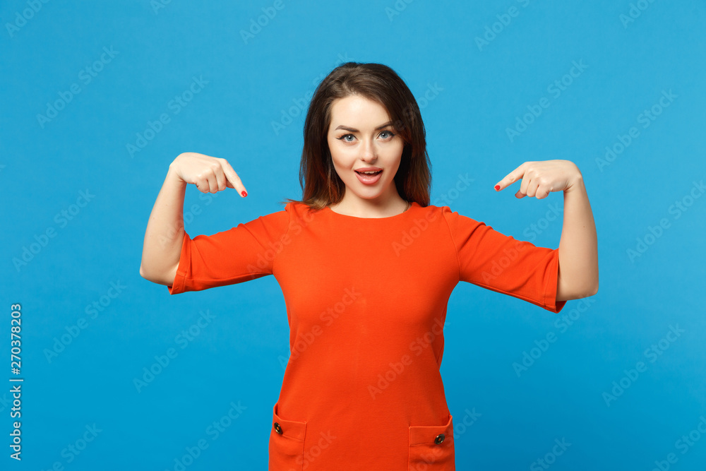 Fototapeta premium Beautiful young woman in red orange dress standing posing pointing hands fingers on workspace isolated over trendy blue wall background, studio portrait. People lifestyle concept. Mock up copy space.