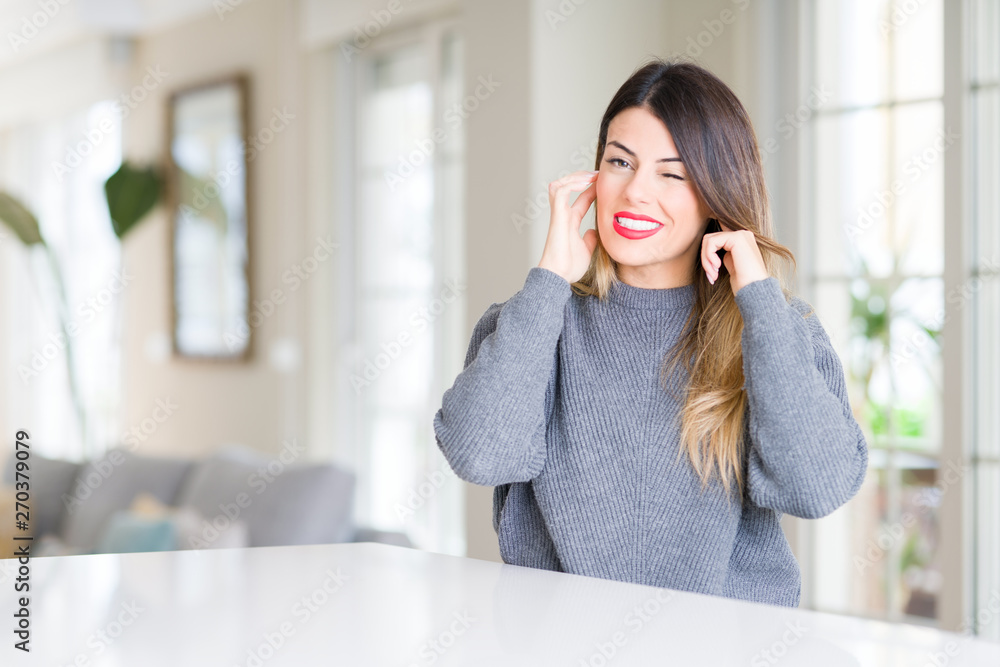 Young beautiful woman wearing winter sweater at home covering ears with fingers with annoyed expression for the noise of loud music. Deaf concept. Young beautiful woman wearing winter sweater at home covering ears with fingers with annoyed expression for the noise of loud music. Deaf concept.