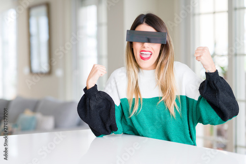 Young beautiful woman blindfold eyes with black censor band at home very happy and excited doing winner gesture with arms raised, smiling and screaming for success. Celebration concept. © Krakenimages.com