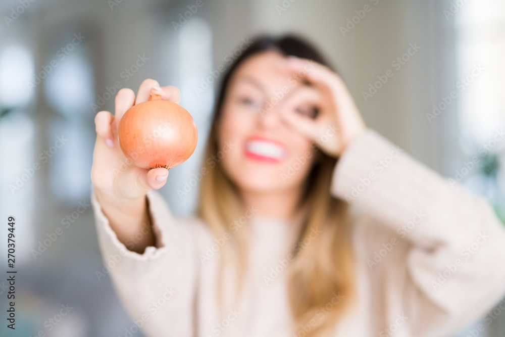 Young beautiful woman holding fresh onion at home with happy face smiling doing ok sign with hand on eye looking through fingers Young beautiful woman holding fresh onion at home with happy face smiling doing ok sign with hand on eye looking through fingers