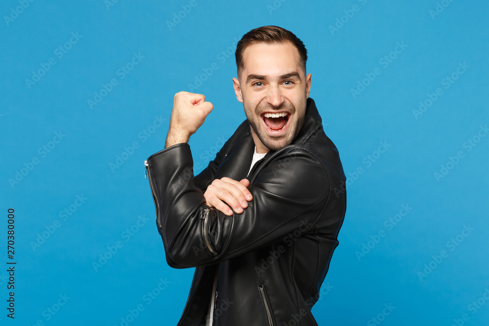 Stylish young bearded man in black leather jacket white t-shirt showing biceps, muscles isolated on blue wall background studio portrait. People sincere emotions lifestyle concept. Mock up copy space. Stylish young bearded man in black leather jacket white t-shirt showing biceps, muscles isolated on blue wall background studio portrait. People sincere emotions lifestyle concept. Mock up copy space.