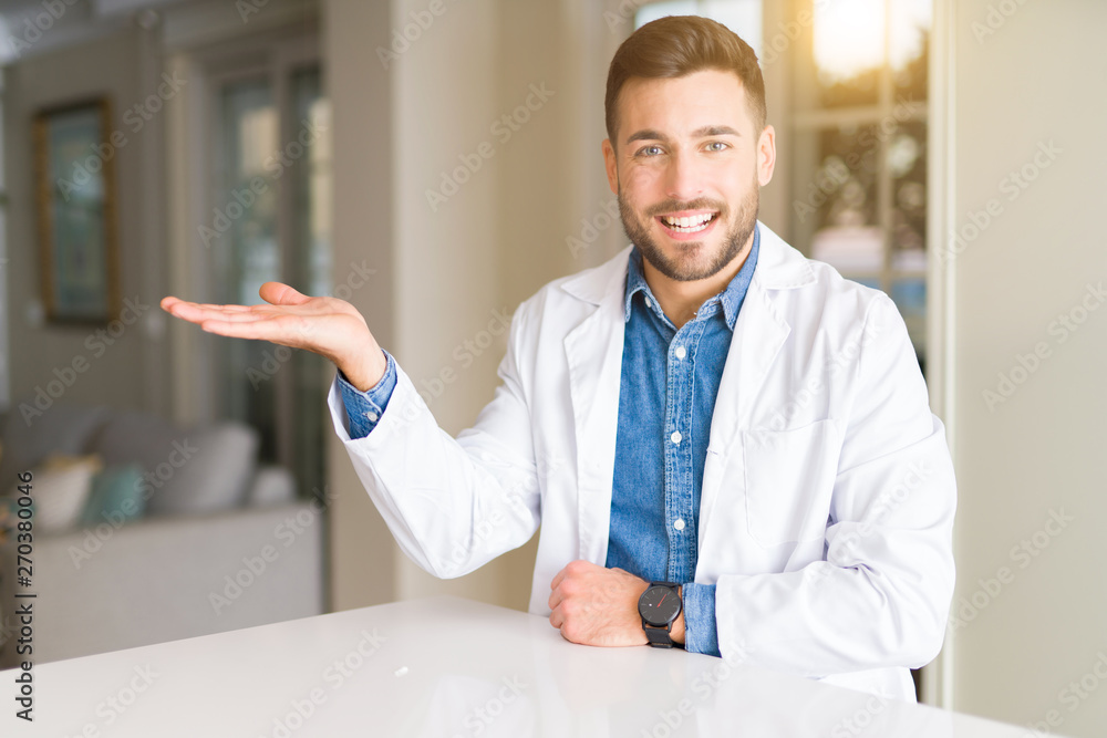 Young handsome doctor man at the clinic smiling cheerful presenting and pointing with palm of hand looking at the camera.