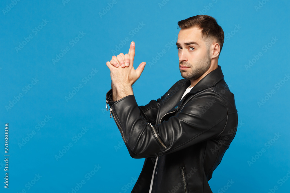 Handsome stylish young unshaven man in black leather jacket white t-shirt holding hands like gun, side view isolated on blue background studio portrait. People lifestyle concept. Mock up copy space Handsome stylish young unshaven man in black leather jacket white t-shirt holding hands like gun, side view isolated on blue background studio portrait. People lifestyle concept. Mock up copy space