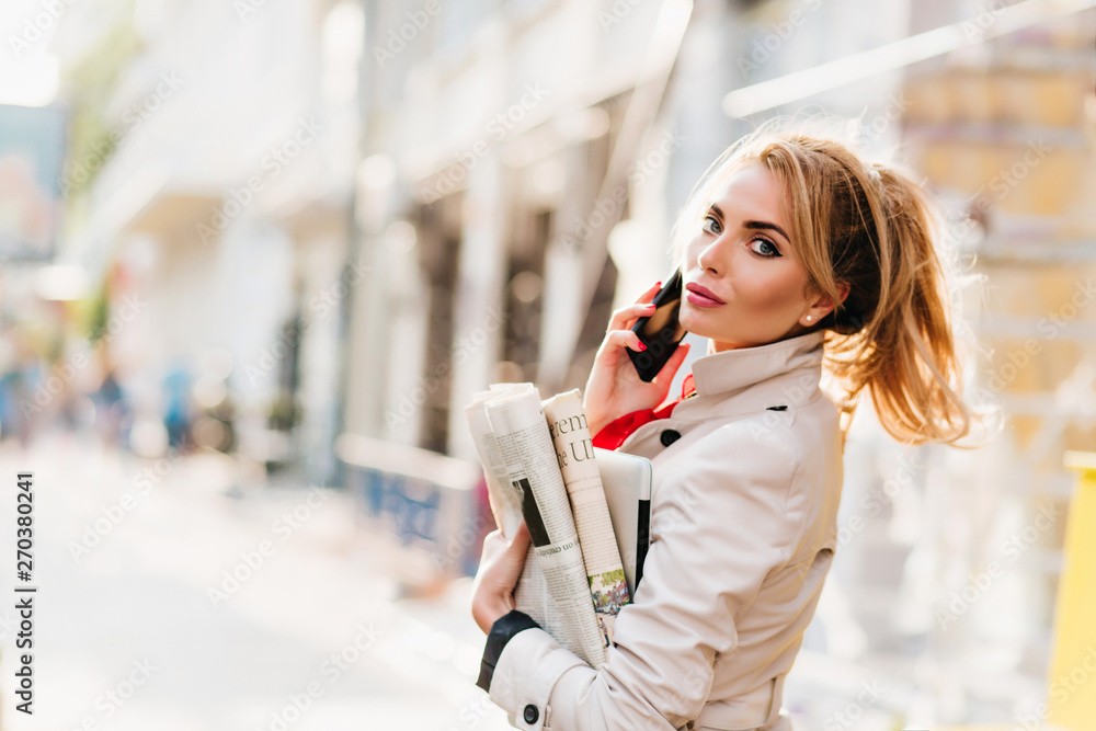 Cheerful fair-haired girl with bright make-up carrying paper and laptop returning home from office. Outdoor portrait of pretty business-lady in beige attire looking around in sunny morning. Cheerful fair-haired girl with bright make-up carrying paper and laptop returning home from office. Outdoor portrait of pretty business-lady in beige attire looking around in sunny morning.