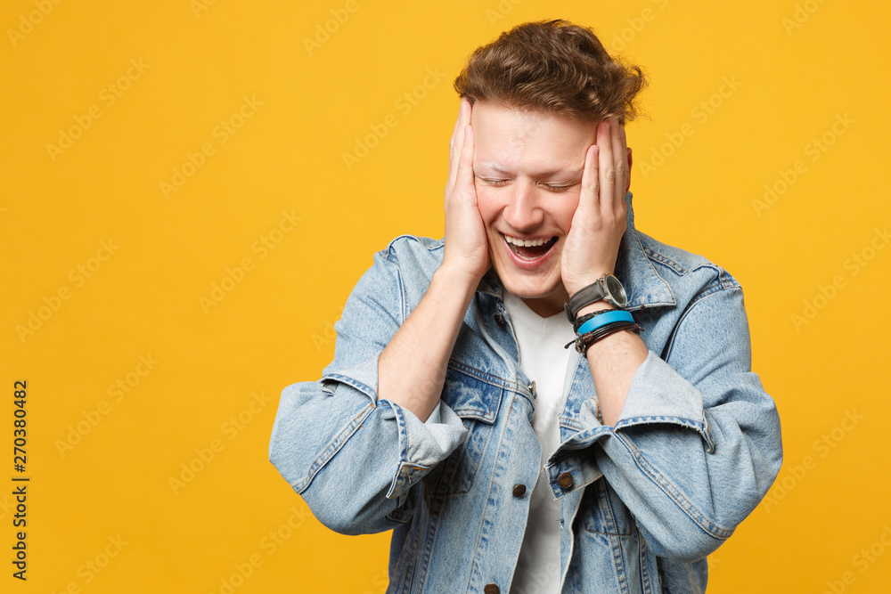 Portrait of laughing young man in denim casual clothes keeping eyes closed, putting hands on head isolated on yellow orange background. People sincere emotions lifestyle concept. Mock up copy space. Portrait of laughing young man in denim casual clothes keeping eyes closed, putting hands on head isolated on yellow orange background. People sincere emotions lifestyle concept. Mock up copy space.