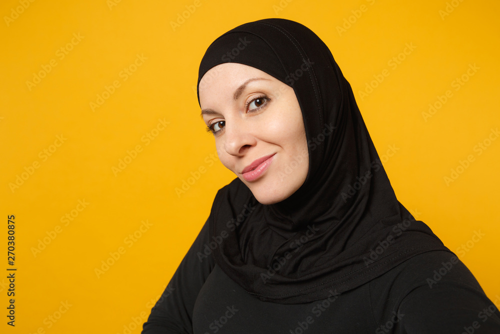 Close up young arabian muslim woman in hijab black clothes doing selfie shot on mobile phone isolated on yellow wall background studio portrait. People religious lifestyle concept. Mock up copy space. Close up young arabian muslim woman in hijab black clothes doing selfie shot on mobile phone isolated on yellow wall background studio portrait. People religious lifestyle concept. Mock up copy space.