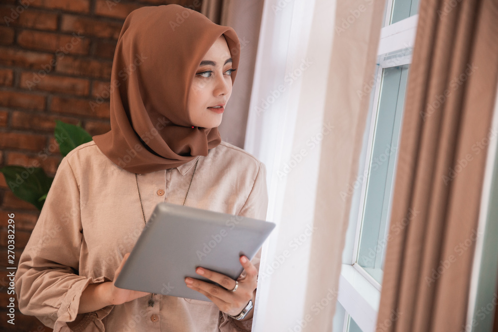 young beautiful woman veiled holding tablet and look out of windows standing near curtains windows in the house young beautiful woman veiled holding tablet and look out of windows standing near curtains windows in the house
