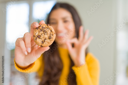 Young woman eating chocolate chips cookies at home doing ok sign with fingers  excellent symbol