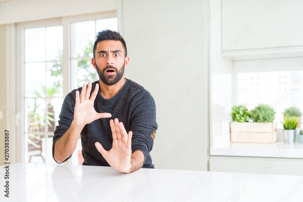 Handsome hispanic man wearing casual sweater at home afraid and terrified with fear expression stop gesture with hands, shouting in shock. Panic concept.
