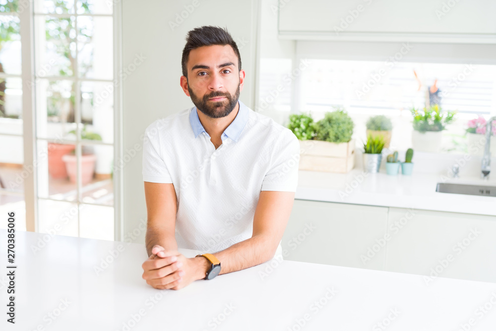 Handsome hispanic man casual white t-shirt at home with serious expression on face. Simple and natural looking at the camera.