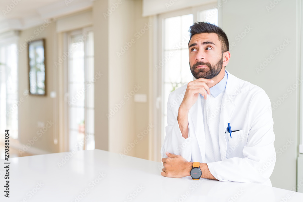 Handsome hispanic doctor or therapist man wearing medical coat at the clinic with hand on chin thinking about question, pensive expression. Smiling with thoughtful face. Doubt concept.