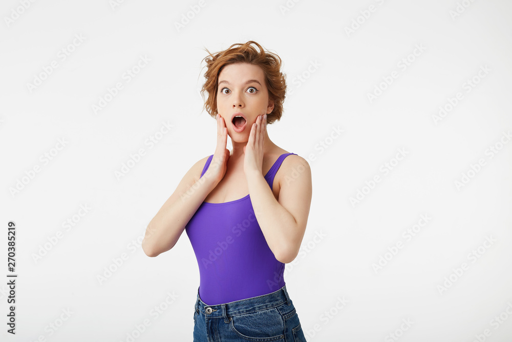 Portrait of amazed young attractive short-haired girl, widely opened her mouth in surprise, touching her face with her hands, stands over white wall. Portrait of amazed young attractive short-haired girl, widely opened her mouth in surprise, touching her face with her hands, stands over white wall.