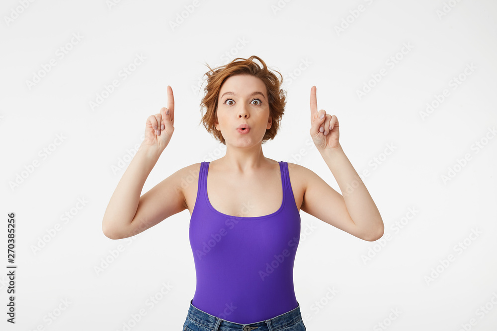 Photo of happy amazed young attractive short-haired girl, opened her mouth in surprise, pointing up at the copy space, stands over white wall. Photo of happy amazed young attractive short-haired girl, opened her mouth in surprise, pointing up at the copy space, stands over white wall.