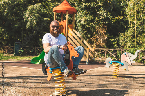 Playful crazy man (dad) riding wooden rocking horse in a park - adult guy (hipster with beard) having fun on a playground
