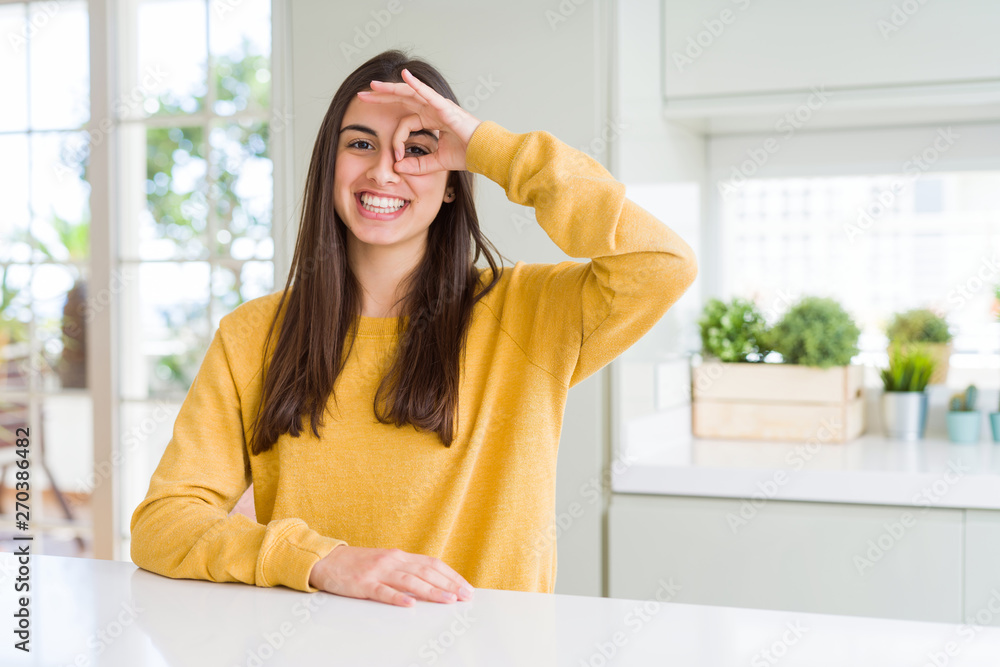 Beautiful young woman wearing yellow sweater doing ok gesture with hand smiling, eye looking through fingers with happy face. Beautiful young woman wearing yellow sweater doing ok gesture with hand smiling, eye looking through fingers with happy face.