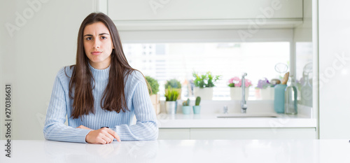 Wide angle picture of beautiful young woman sitting on white table at home skeptic and nervous, frowning upset because of problem. Negative person.
