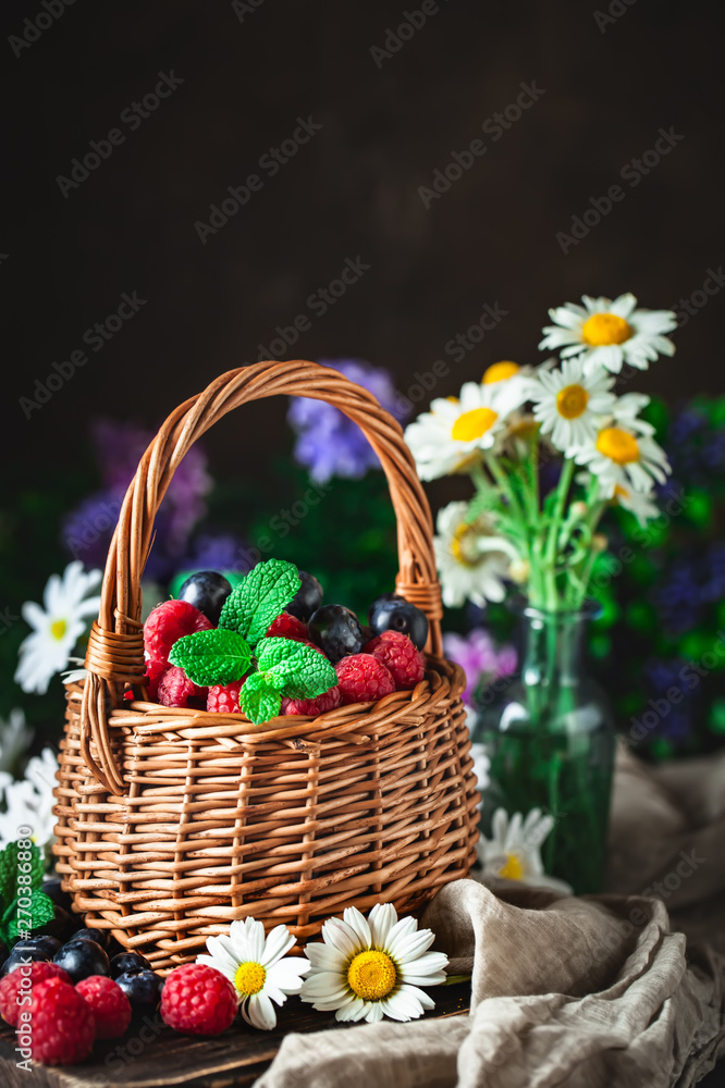 Raspberries and blueberries in a basket with chamomile and leaves on a dark background. Summer and healthy food concept. Selective focus. Background with copy space.