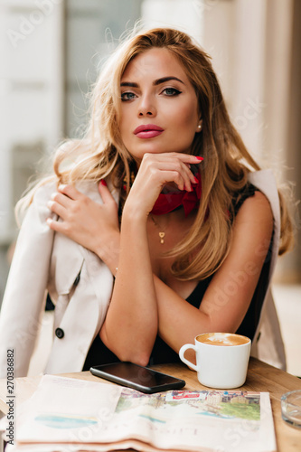 Lightly-tanned fashionable girl sitting at the table with phone and cup of coffee on it. Portrait of lovely blonde girl in beige coat spending free time in favorite cafe.