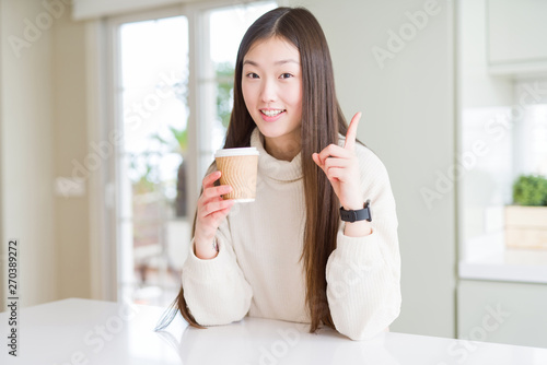 Beautiful Asian woman drinking a coffee in a take away paper cup surprised with an idea or question pointing finger with happy face, number one