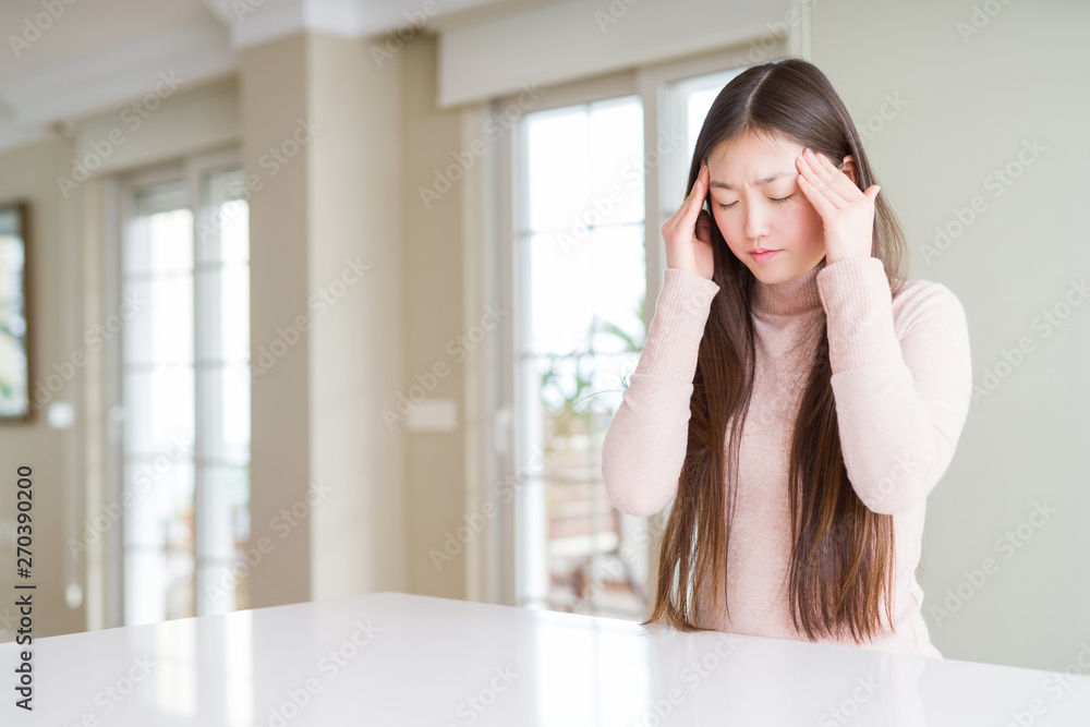 Beautiful Asian woman wearing casual sweater on white table with hand on head for pain in head because stress. Suffering migraine. Beautiful Asian woman wearing casual sweater on white table with hand on head for pain in head because stress. Suffering migraine.