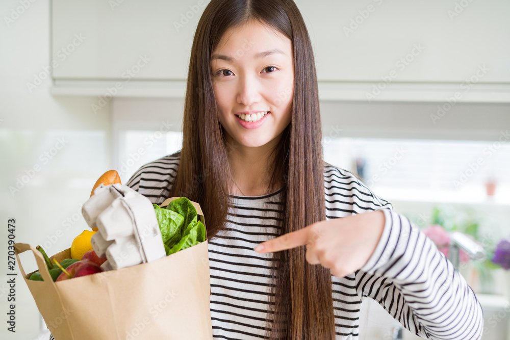 Beautiful Asian woman holding paper bag of fresh groceries very happy pointing with hand and finger