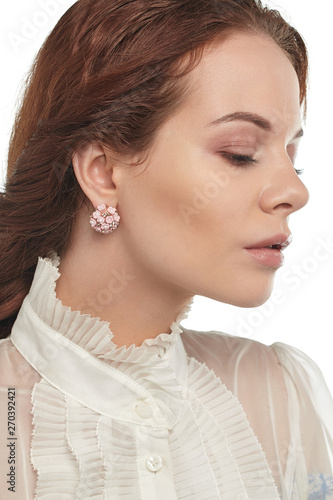 Portrait of girl with brown hair, wearing white blouse with ruffles, looking down on white background. The lady is wearing massive earrings, adorned with crystals and pale pink floral application.