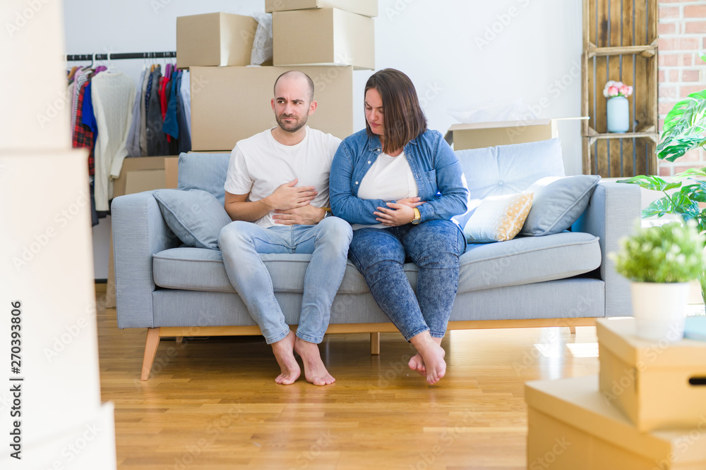 Young couple sitting on the sofa arround cardboard boxes moving to a new house with hand on stomach because nausea, painful disease feeling unwell. Ache concept. Young couple sitting on the sofa arround cardboard boxes moving to a new house with hand on stomach because nausea, painful disease feeling unwell. Ache concept.