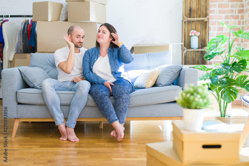 Young couple sitting on the sofa arround cardboard boxes moving to a new house smiling with hand over ear listening an hearing to rumor or gossip. Deafness concept. Young couple sitting on the sofa arround cardboard boxes moving to a new house smiling with hand over ear listening an hearing to rumor or gossip. Deafness concept.