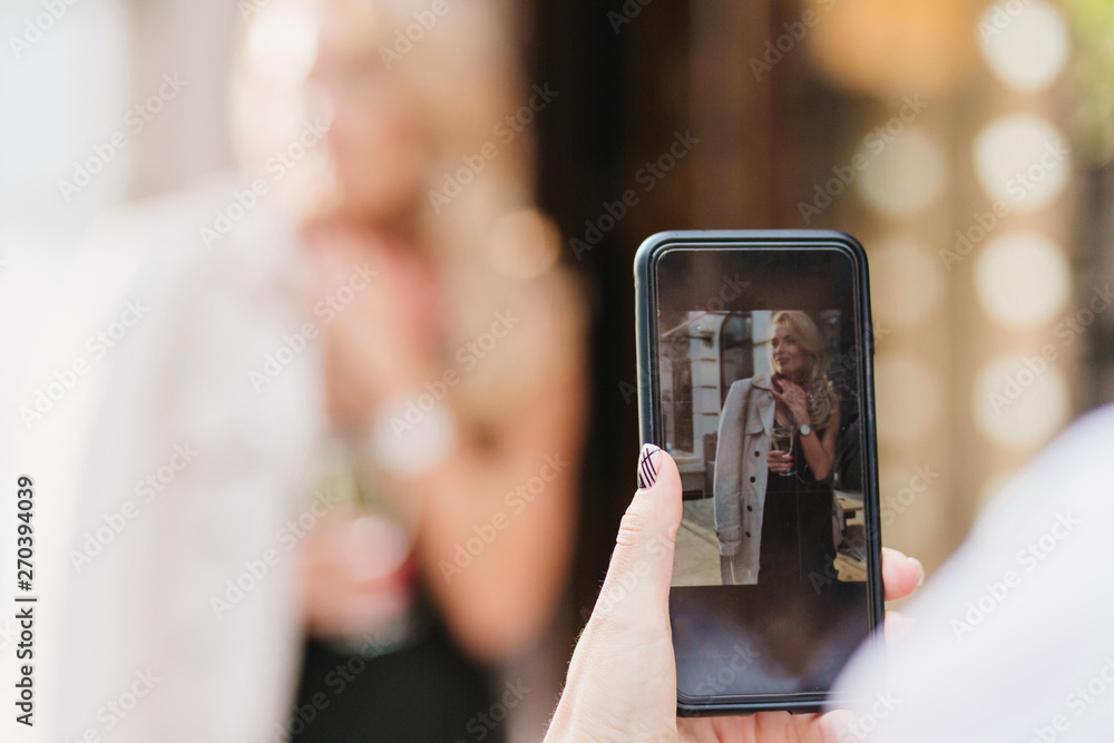 Girl with elegant manicure taking picture of charming female friend which posing outdoor. Girl holding smartphone with photo of graceful woman at screen.