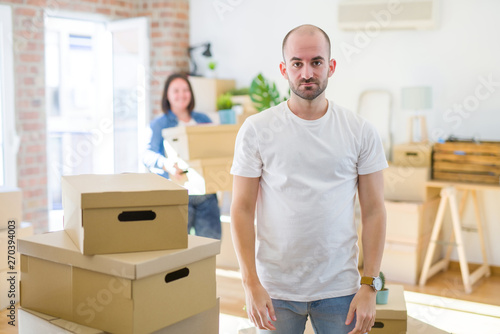 Young couple arround cardboard boxes moving to a new house, bald man standing at home with serious expression on face. Simple and natural looking at the camera.