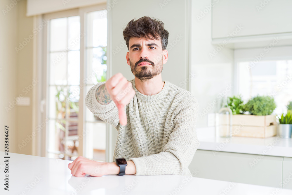Young man wearing casual sweater sitting on white table looking unhappy and angry showing rejection and negative with thumbs down gesture. Bad expression.