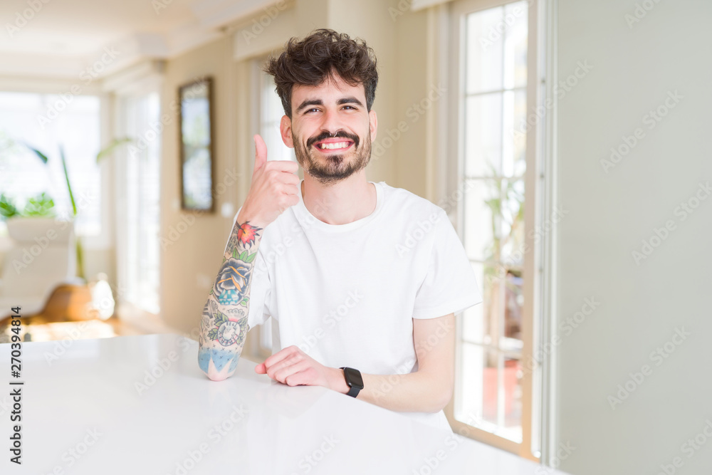 Fototapeta premium Young man wearing casual t-shirt sitting on white table doing happy thumbs up gesture with hand. Approving expression looking at the camera showing success.