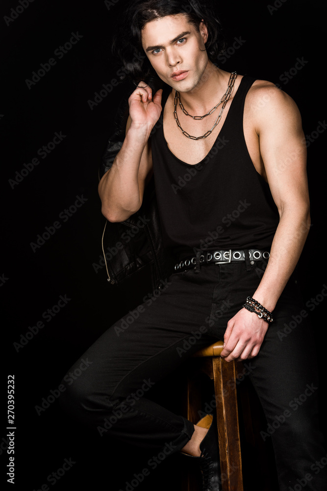 handsome man holding leather jacket and looking at camera while sitting on chair isolated on black handsome man holding leather jacket and looking at camera while sitting on chair isolated on black