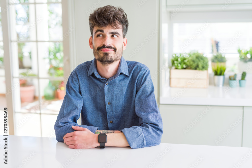 Young man wearing casual shirt sitting on white table smiling looking side and staring away thinking. Young man wearing casual shirt sitting on white table smiling looking side and staring away thinking.