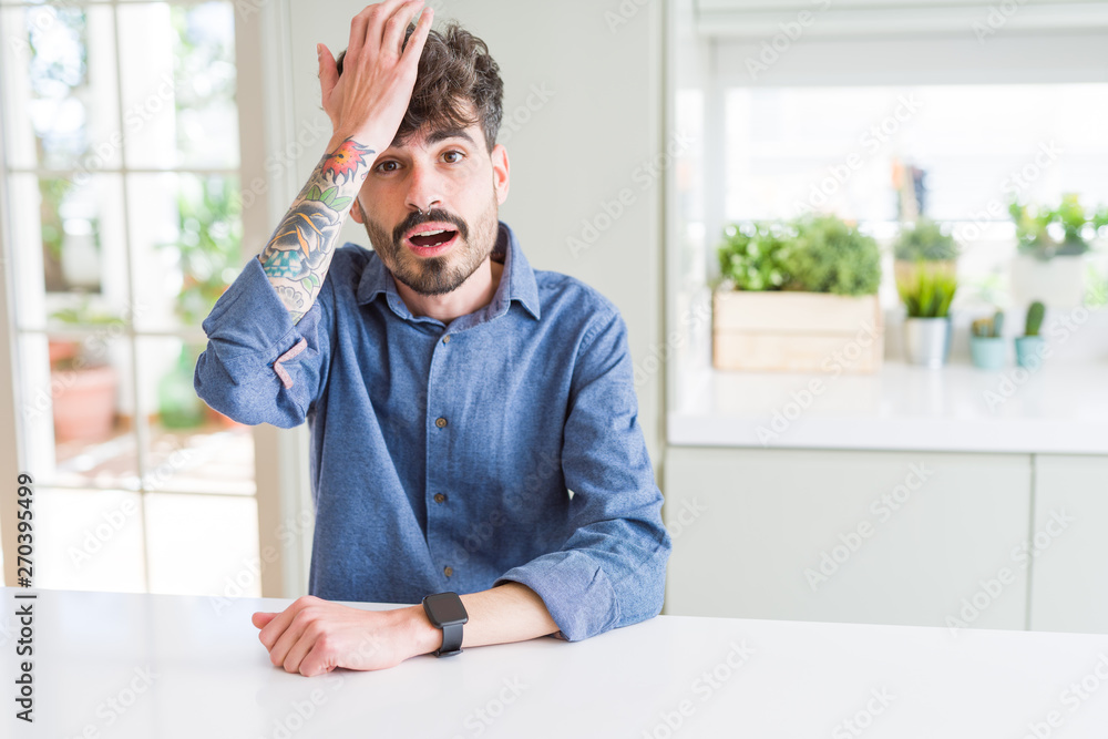 Young man wearing casual shirt sitting on white table surprised with hand on head for mistake, remember error. Forgot, bad memory concept. Young man wearing casual shirt sitting on white table surprised with hand on head for mistake, remember error. Forgot, bad memory concept.