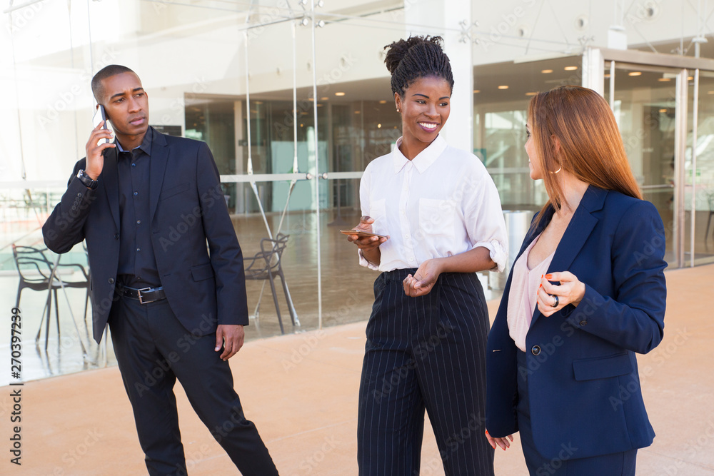 Positive ladies discussing project while walking over corridor. Curious black businessman listening to conversations of women while calling on mobile phone. Business company staff concept Positive ladies discussing project while walking over corridor. Curious black businessman listening to conversations of women while calling on mobile phone. Business company staff concept