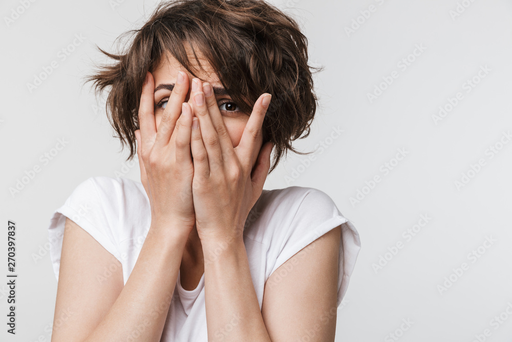 Photo of frightened woman with short brown hair in basic t-shirt keeping hands over her face and looking at camera Photo of frightened woman with short brown hair in basic t-shirt keeping hands over her face and looking at camera