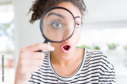 Young african american woman looking through magnifying glass scared in shock with a surprise face, afraid and excited with fear expression