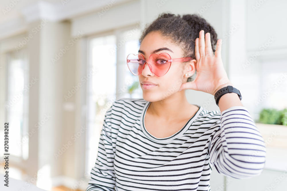 Beautiful young african american woman with afro hair wearing crazy pink heart glasses smiling with hand over ear listening an hearing to rumor or gossip. Deafness concept. Beautiful young african american woman with afro hair wearing crazy pink heart glasses smiling with hand over ear listening an hearing to rumor or gossip. Deafness concept.