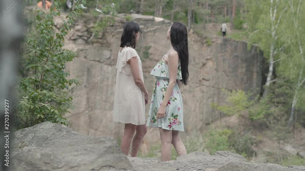 Portrait two beautiful women in short dresses standing on rocky ground with wild flowers and looking forest enjoying nature. Adorable girlfriends spending the weekend together walking outdoors. Portrait two beautiful women in short dresses standing on rocky ground with wild flowers and looking forest enjoying nature. Adorable girlfriends spending the weekend together walking outdoors.