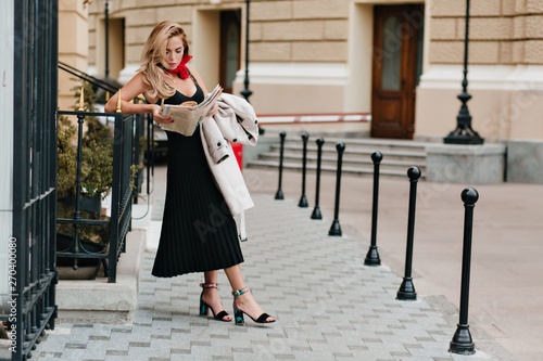 фотографія Fascinating fair-haired woman wears colorful sandals reading newspaper leaning to iron fence