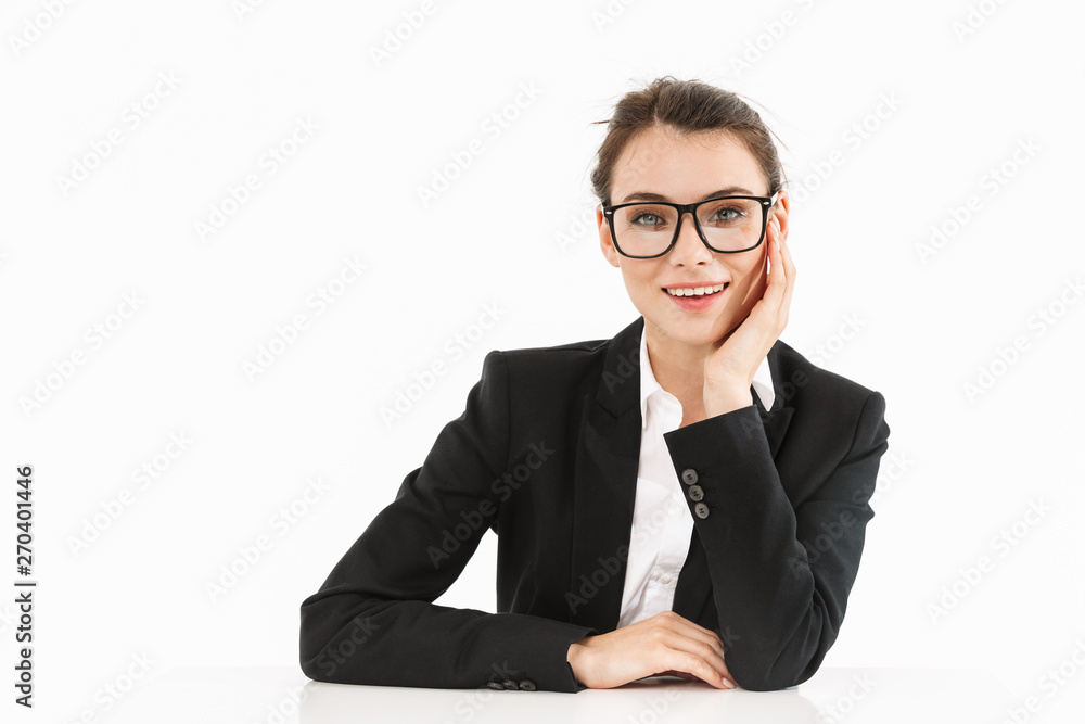 Photo of smiling female worker businesswoman dressed in formal wear looking at camera while working and sitting at desk in office Photo of smiling female worker businesswoman dressed in formal wear looking at camera while working and sitting at desk in office