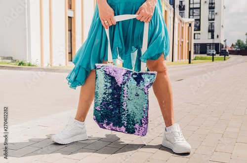 Fashionable stylish woman in white sneakers, holding a trend backpack-bag of two-sided blue and purple booze in a city street.